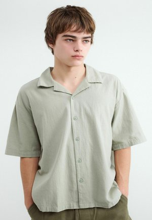 Jeune homme aux cheveux châtain clair portant une chemise ample vert pâle à manches courtes boutonnée, les mains dans les poches, regardant légèrement vers la gauche.