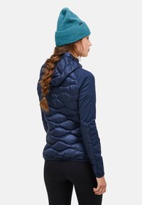 Marinefarbene isolierte Jacke mit gestepptem Design, mit Kapuze und einfarbigen Ärmeln, kombiniert mit einer türkisfarbenen Strickmütze.