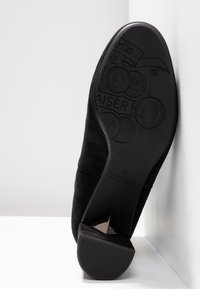 Chaussure à talon haut en suède noir avec un bout pointu et une semelle en caoutchouc texturé, présentant des motifs de pièces en relief au fond.