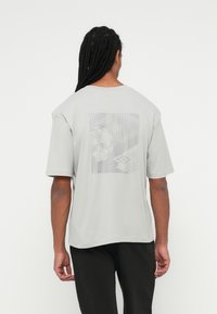 Homem com longas tranças vestido com uma t-shirt cinza clara com um discreto design de bola de futebol geométrica e padrão nas costas, combinado com calças pretas.