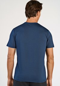 Mand med kort brunt hår, der vender væk, iført en ensfarvet marineblå T-shirt med korte ærmer og mørke bukser mod en hvid baggrund.
