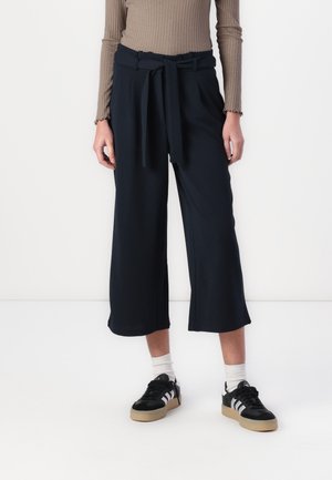 Person mit schwarzen, weit geschnittenen, cropped Pants mit Gürtel, beige-rippigem, langärmligem Oberteil, weißen Socken und schwarzen Sneakers mit weißen Streifen.
