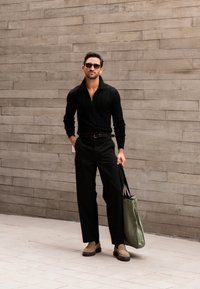Suéter negro con relieve y media cremallera, pantalones negros, mocasines de ante color marrón claro y un bolso verde, contra una pared gris texturizada.