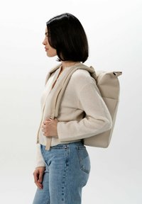 Beige rolltop rugzak met verstelbare schouderbanden, een gladde textuur en een zijzak, gedragen over een licht sweater en denim jeans.