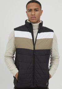 Padded Weste in Schwarz, Beige und Weiß. Mit hohem Kragen, Reißverschluss und strukturiertem Strickrollkragen darunter. Farbblöcke im Kontrast.