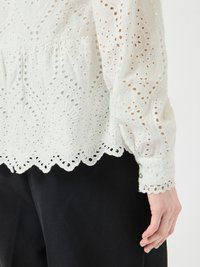 Blusa bianca ricamata con motivi floreali perforati, orlo scallopato e polsini con bottoni, abbinata a pantaloni scuri.