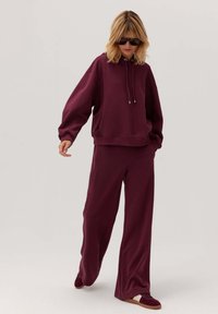 Burgundowa bluza z kapturem i sznurkami, w luźnym kroju, zestawiona z szerokimi spodniami. Oba elementy wykonane z miękkiego materiału, zapewniającego swobodną sylwetkę.