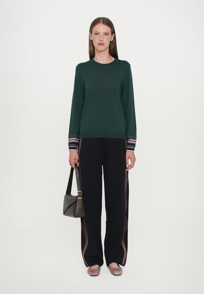 Pulover tricotat verde închis cu manșete cu model, asortat cu pantaloni negri cu dungi colorate pe lateral. Completat cu o poșetă gri și pantofi plata multicolori.