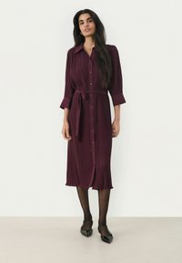Burgundy-harmaatikkojakaan mekko, jossa on nappisalpa etuosassa, pitkät hihat ja sidottu vyö. Mekossa on polvien alapuolella ulottuva pituus ja teksturoitu kangas.