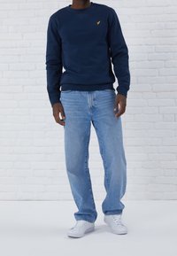 Mörkblå tröja med gult logotyp, i kombination med ljusblå raka jeans och vita sneakers. Mot en vit tegelvägg.