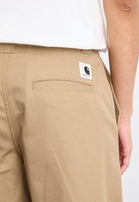Pantaloni in cotone beige con una tasca posteriore che presenta un'etichetta bianca con un logo nero. Il tessuto ha una consistenza liscia ed è sagomato.