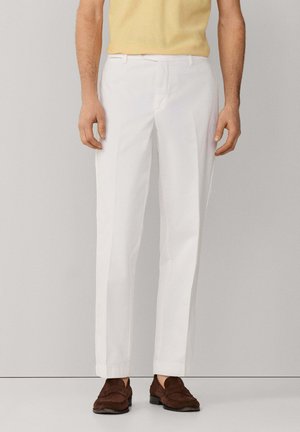 Man met slanke witte broek, een geel shirt met korte mouwen en bruine suède loafers, staand tegen een effen achtergrond.