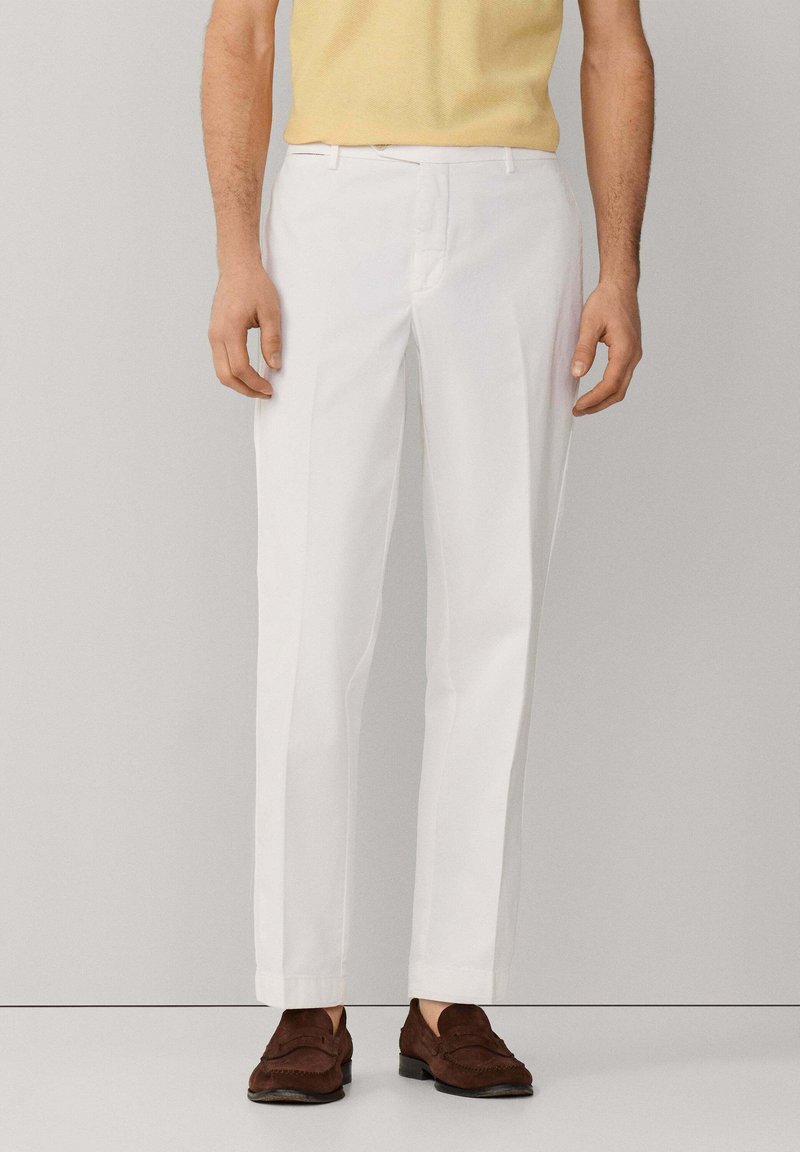 Uomo che indossa pantaloni bianchi attillati, una camicia gialla a maniche corte e mocassini marroni in suede, in piedi davanti a uno sfondo neutro.