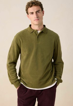Jonge man met krullend haar, gekleed in een olijfgroen overhemd met lange mouwen, een wit onderhemd en donker bordeauxrode corduroy broek, met zijn handen in zijn zakken.