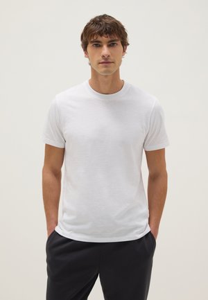 Junger Mann mit kurzem braunem Haar, trägt ein schlichtes weißes T-Shirt mit kurzen Ärmeln und schwarze Hosen, steht mit den Händen in den Taschen.