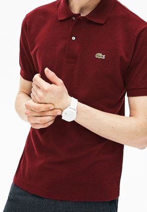 Homme portant un polo bordeaux avec un logo de crocodile et une montre blanche au poignet, ajustant son poignet gauche avec sa main droite.