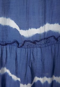 Modrá a bílá látka se vzorem zahrnujícím vlnitý tie-dye design s volánkovým lemem a nabranou texturou podél švu.