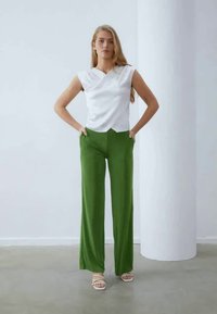 White top with a V neckline and cap sleeves paired with high-waisted, wide-leg green pants in a smooth fabric. Model stands with hands on hips.