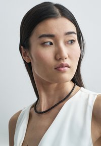 Μαύρο κολιέ με χάντρες και λεία υφή, φορεμένο με λευκό ρούχο. Το μοντέλο έχει ίσια μαλλιά και είναι ελαφρώς στραμμένο προς τη μία πλευρά.