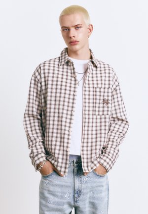 Ung person med kort blondt hår iført beige og brunternet skjorte over hvid t-shirt og lyseblå jeans med hvid broderi, hænderne i lommerne.