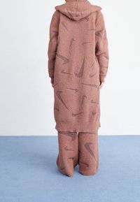 Överdimensionerad, rosa-brun fleecehuva med en strukturerad yta, med ett subtilt brunt Nike-logomönster både på toppen och de matchande byxorna.