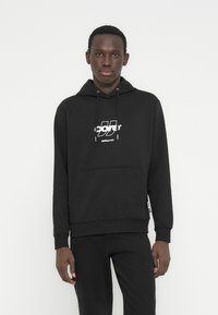 Νεαρός άνδρας που φοράει μαύρο φούτερ Jack & Jones και μαύρο παντελόνι, στέκεται μπροστά σε απλό λευκό φόντο.