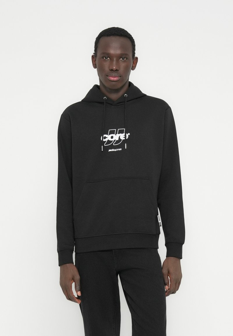 Νεαρός άνδρας που φοράει μαύρο φούτερ Jack & Jones και μαύρο παντελόνι, στέκεται μπροστά σε απλό λευκό φόντο.