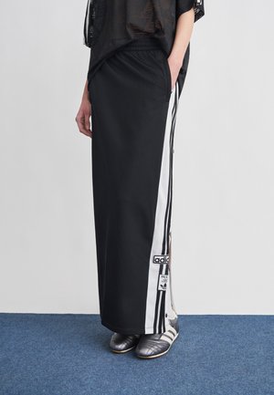 Oseba oblečena v dolgo črno krilo z belimi stranskimi črtami in logotipom Adidas, obuta v črno-bele Adidas superge, stoji na modrem preprogi.