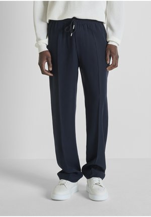 Uomo con pantaloni blu navy con coulisse, maglione bianco e sneakers bianche, in piedi davanti a uno sfondo grigio chiaro uniforme.