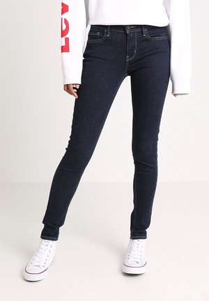 Personne portant un jean skinny bleu foncé, des baskets montantes blanches, et un haut blanc à manches longues avec un lettrage rouge sur la manche.