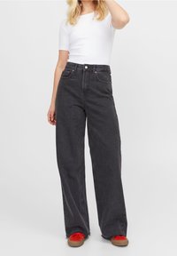 Svarta denimjeans med vida ben och hög midja, med orangea sömmar, två framfickor och knappstängning. Matchat med en vit topp.