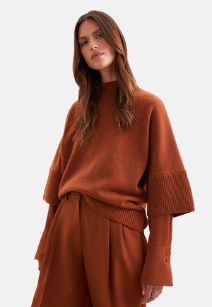 Femme aux longs cheveux bruns portant un pull en maille ample couleur rouille sur un pantalon large assorti de couleur rouille sur un fond uni.