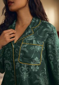 Zelená pyžamová blúzka s kvetinovým vzorom a žltým lemovaním. Má náprsné vrecko a klopový golier s hladkým povrchom.