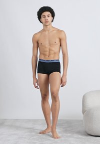 Cuecas boxer pretas com cintura azul, feitas de tecido macio, exibidas em um modelo masculino em pé contra um fundo claro.