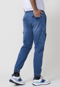Modre denim hlače s cargo krojem in elastičnim pasom, z ozkimi nogami ter več žepi. Ponašajo se z gladko teksturo in lahko zasnovo.