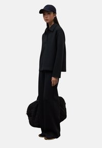 Mujer joven vestida con una blusa negra de manga larga, pantalones de pierna ancha, zapatillas y una gorra negra, sosteniendo un gran bolso negro acolchado frente a un fondo blanco.