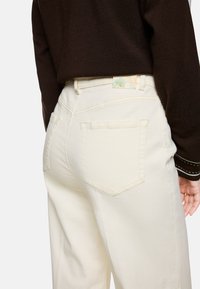 Weiße Denim-Hose mit hoher Taille, die über hinten liegende Taschen verfügt und ein Markenetikett in sanften Farben am Bund sowie minimalistische Nähte aufweist.