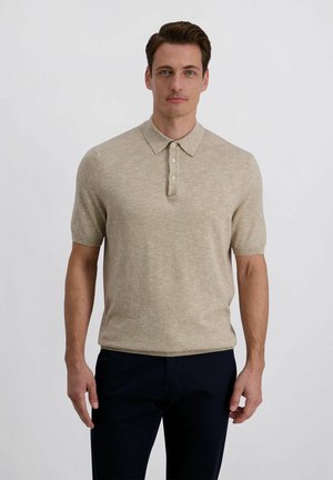 Mann mit kurzen braunen Haaren, der ein beiges, kurzärmeliges Poloshirt und dunkle Hosen trägt, vor einem schlichten hellen Hintergrund stehend.