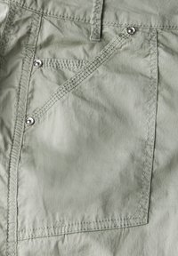 Jasnozielone spodnie z bliska, ukazujące przednią kieszeń z ukośnym potrójnym przeszyciem i trzema metalowymi nitami w pobliżu brzegów kieszeni.