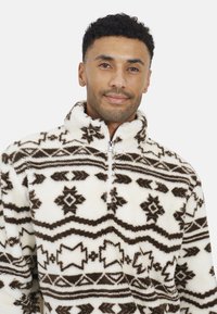 Pullover bianco in pile con un motivo geometrico marrone, dotato di collo alto e zip frontale a metà. La texture morbida aggiunge calore.