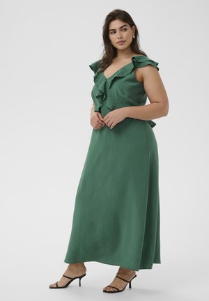 Femme portant une robe maxi verte sans manches avec un col à volants et des sandales à talons noires, debout devant un fond clair uni.