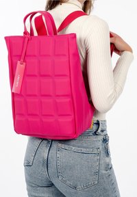 Mochila acolchada rosa con dos asas superiores y correas ajustables, hecha de material texturizado. Incluye una etiqueta de marca en el lateral.