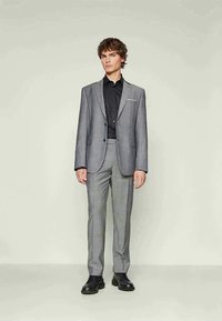 Young man standing in a gray checkered suit, black dress shirt, and black shoes against a plain light background.