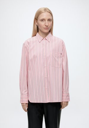 Femme blonde portant une chemise rose ample à boutons avec de fines rayures verticales et un pantalon noir, debout devant un fond blanc uni.