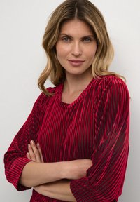 Blusa a righe rosse e nere con texture a coste; presenta un collo rotondo e maniche a tre quarti con dettagli arricciati.
