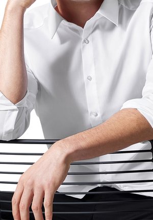 Mężczyzna w białej koszuli na guziki siedzi tyłem na czarnej metalowej krześle, z jedną ręką opartą na oparciu krzesła.