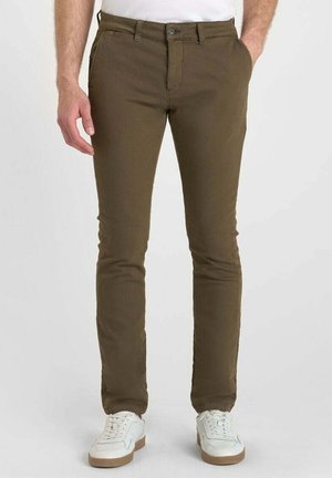 Man in slim-fit bruine broek en witte sneakers, met de handen in de zakken voorop, tegen een effen lichte achtergrond.