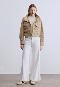 Κοντή μπεζ ζακέτα με δύο τσέπες στο στήθος, κουμπιά κλεισίματος και μεγάλο γιακά, συνδυασμένη με λευκό παντελόνι με φαρδιά πόδια και μπεζ παπούτσια.