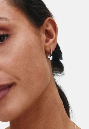 Pendiente de aro plateado con un colgante en forma de corazón. Textura suave, acabado pulido y diseño de cierre simple para un uso seguro.