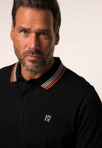 Polo shirt nera con colletto a righe arancioni e bianche. Presenta un piccolo logo bianco sul lato sinistro del petto. Tessuto liscio.